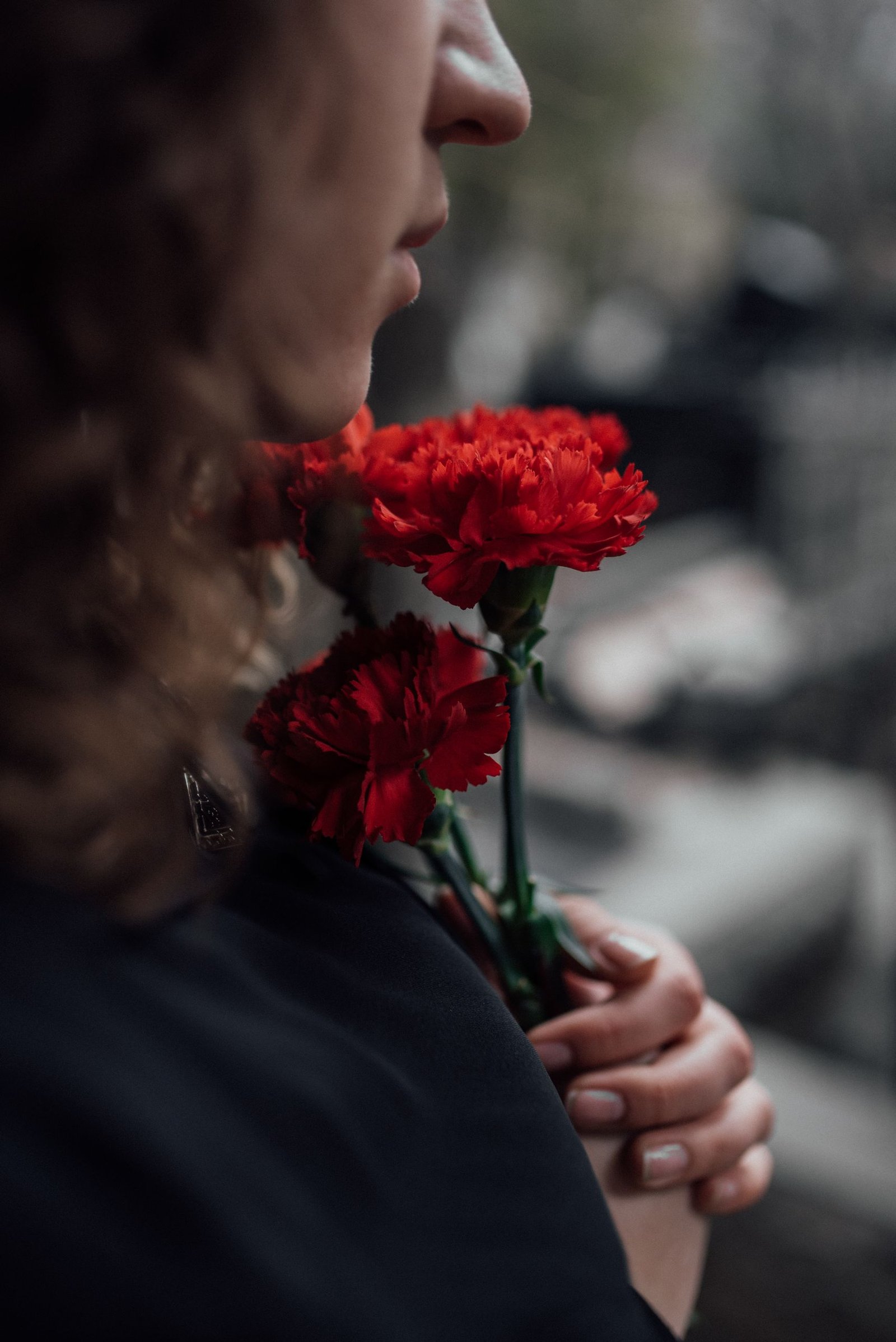 A thoughtful woman holds red carnations, evoking themes of loss and memory in an outdoor setting.