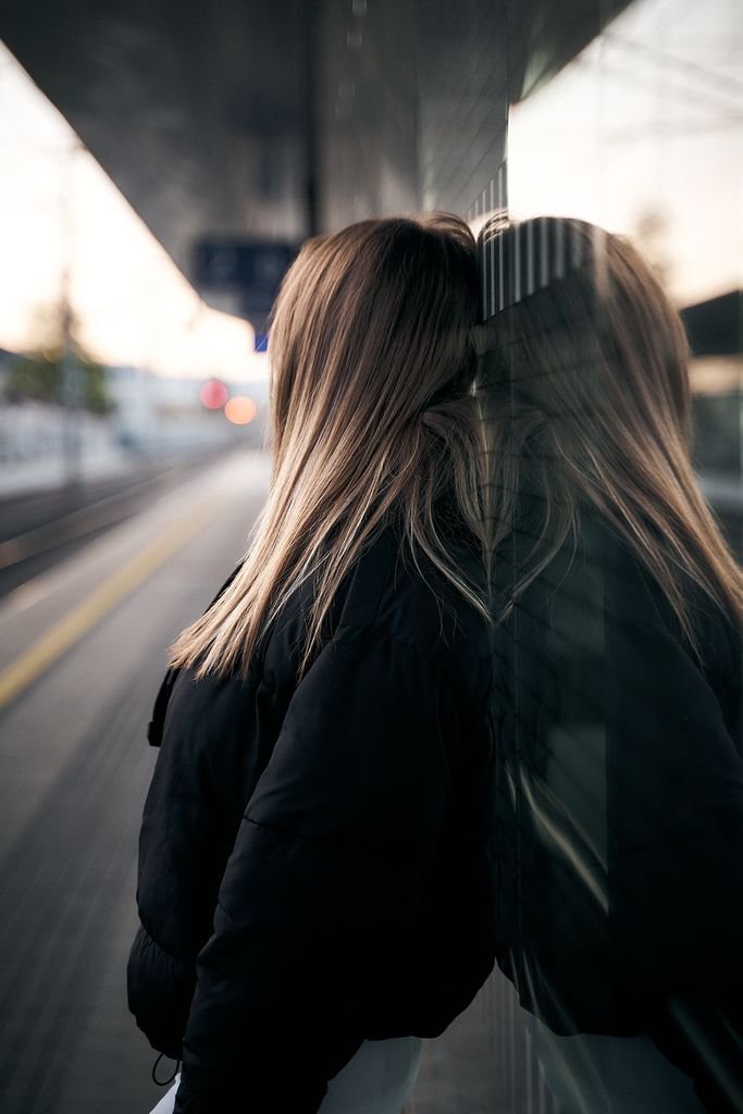 A woman leans against a reflective surface at an Austrian train station, expressing contemplation.